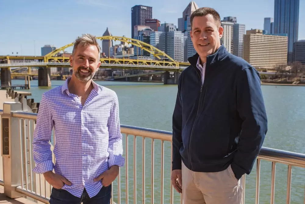 Behr and Rydzewski smile in Pittsburgh’s North Side in front of a yellow bridge on the Allegheny River